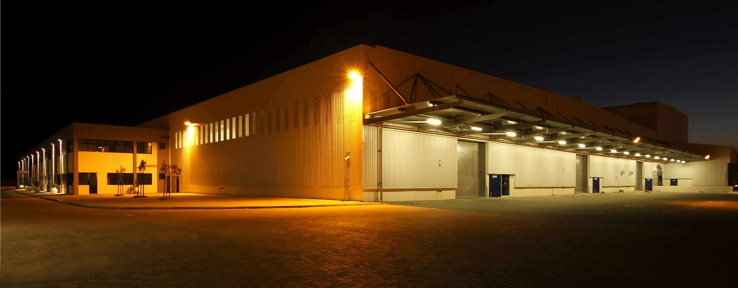 Large commercial warehouse facility at night with bright exterior lighting, loading docks, and secured entry doors, highlighting the need for strong physical security and surveillance systems.